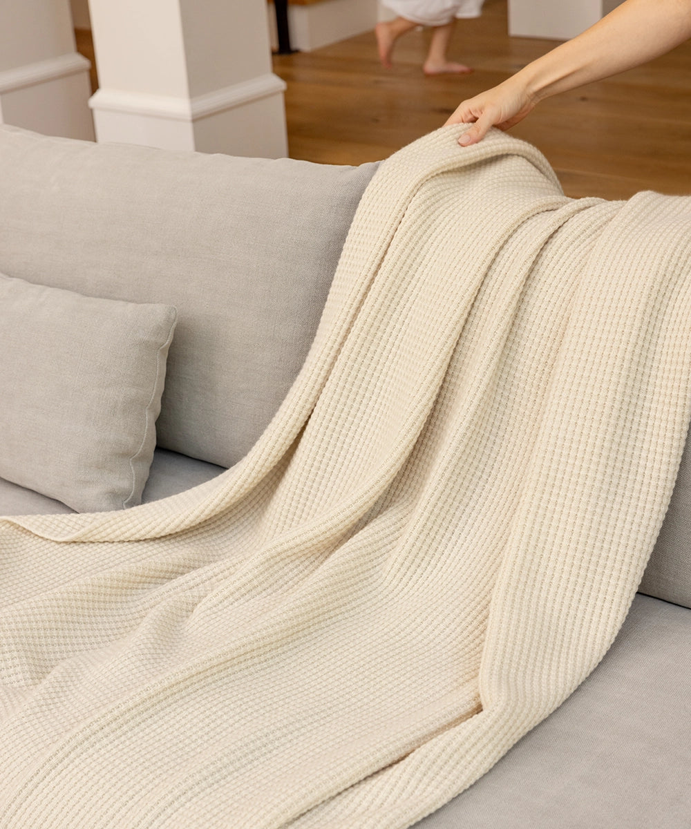 Woman draping a cream AU Baby Popcorn throw blanket over the back of a grey couch while a small child runs around in the background. 