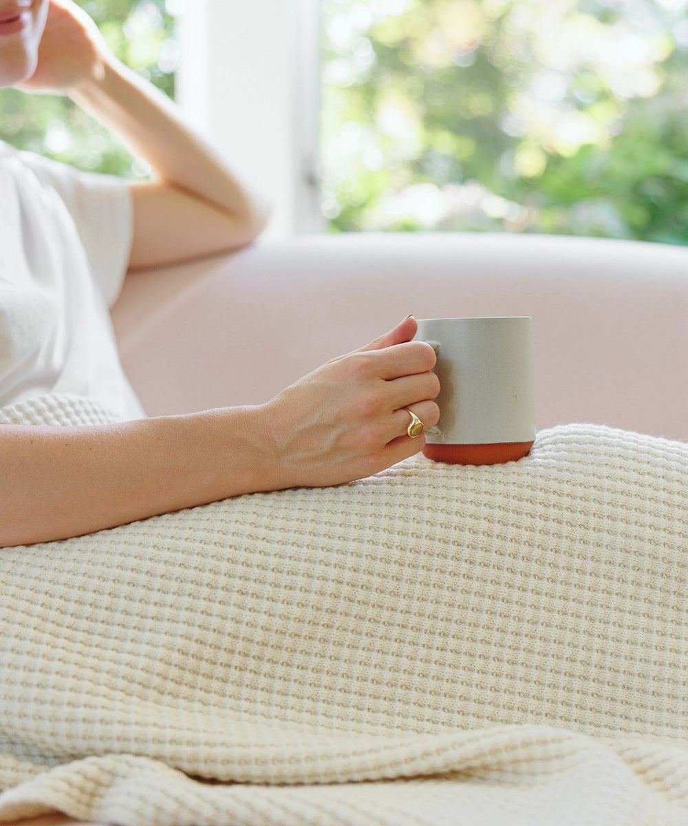 Woman relaxing with coffee, snuggled up with a beautiful cream AU Baby 100% merino throw blanket.